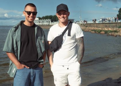 Two young men smiling near the sea on a sunny day, enjoying the beach atmosphere.